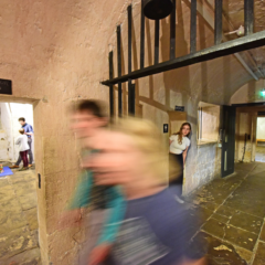 A family plays with toys in the background, two blurred adults walk past the camera, whilst a third adult looks out from a doorway behind them
